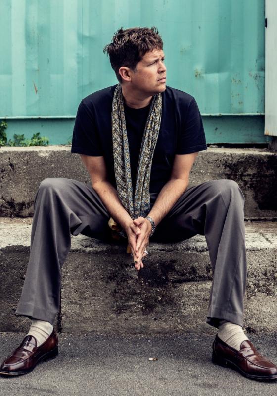 The performer sits on a concrete step wearing black and grey, looking into the distance