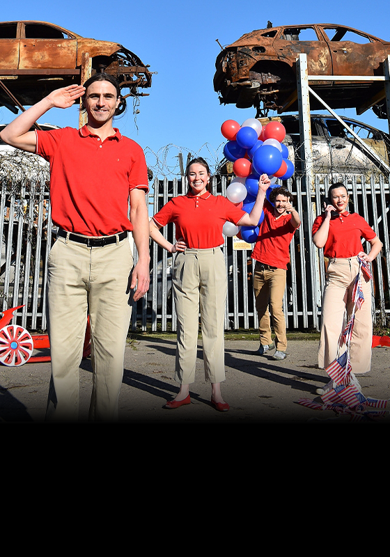 Four performers wearing red polos and khaki pants stand in front of a car junkyard