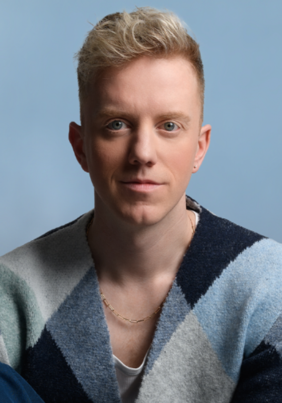 An image of James Barr, looking directly at the camera. They are wearing a blue tone block cardigan and sitting against a blue background