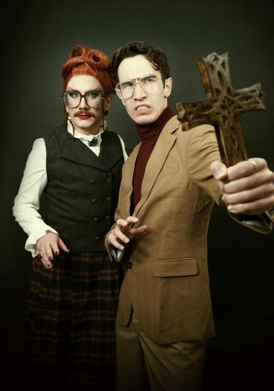Two performers stand in front of a black background. One of them is holding up a wooden crucifix.