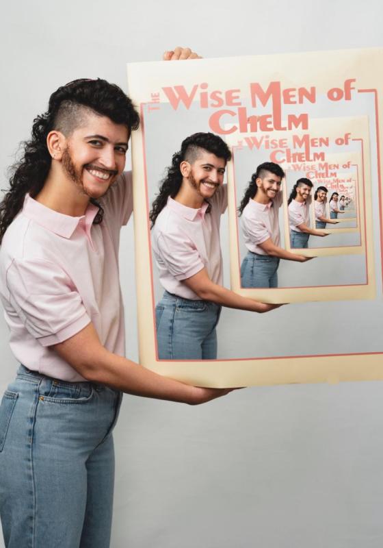 The performer smiles while holding a a mirror which reads 'The Wise Men if Chelm'  in bold red lettering. Their reflection can be repeatedly seen within the mirror.