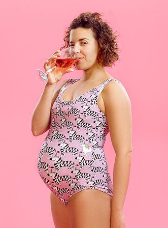 A pregnant woman drinking a glass of wine, looking directly out: she is wearing a pink leotard with zebras & standing in front of a pink background