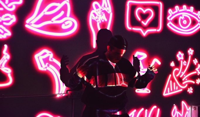 A performer in a cap is lit by projection of neon symbols - hearts, lips, eyes, crossed fingers