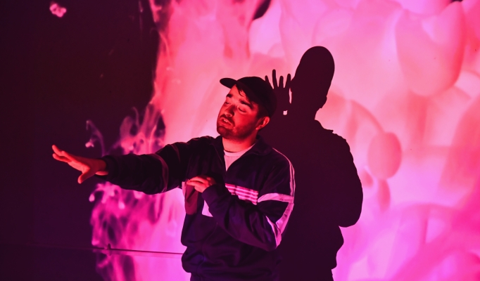 A performer in a cap is lit by swirling pink projection