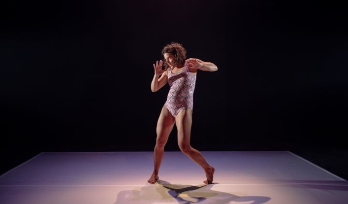 A pregnant woman wearing a pink leotard stands, mid-movement on stage 