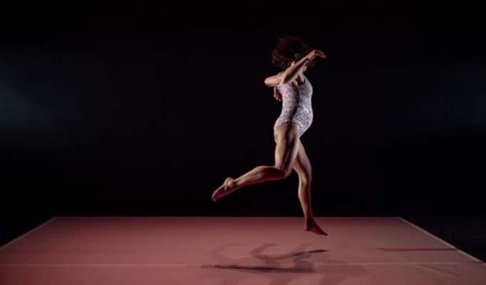 A pregnant woman in a pink leotard is mid-leap in a movement sequence 