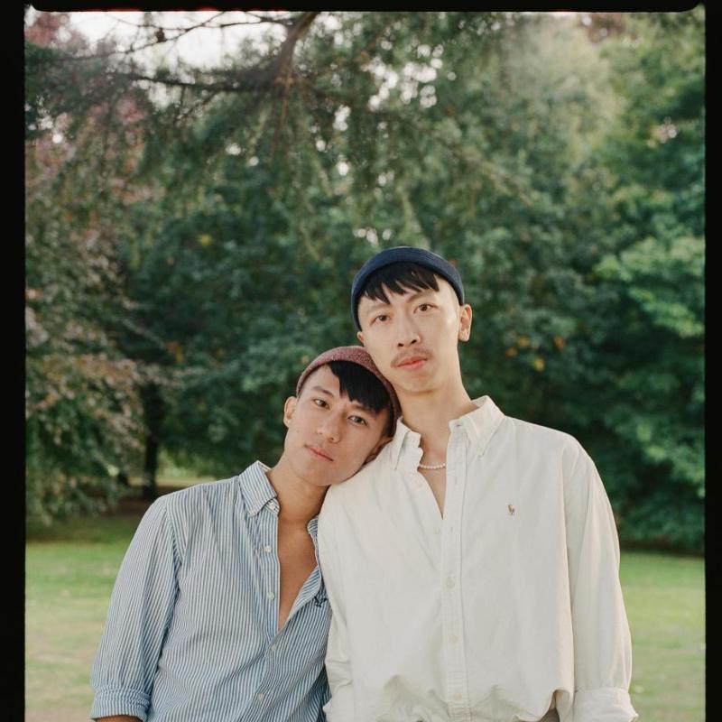2 People standing in in a park. The shorter person leans their head on the shoulder of the other. They both have their hands in their pockets and are wearing shirts and smart trousers.