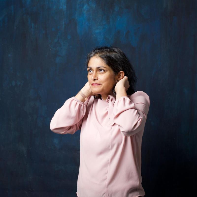 A woman in a pink shirt stands in front of a dark ocean blue backdrop putting her hair behind her ears looking up positively.