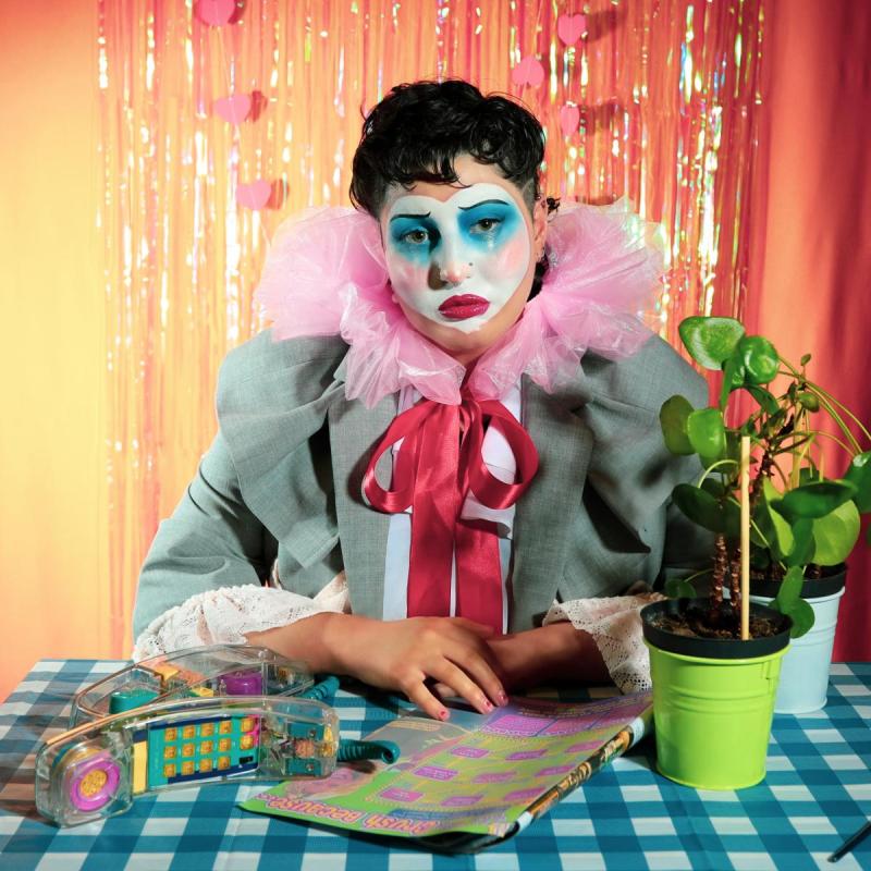 Saffia, a non-binary white person with short curly dark hair, wears a pink clown collar with a large red bow and a grey blazer. They are sitting with their arms on a blue gingham table, showing a plant, a noughties magazine and a transparent telephone with multicoloured keys. The backdrop is sunset orange with iridescent fringe and hanging heart garlands.