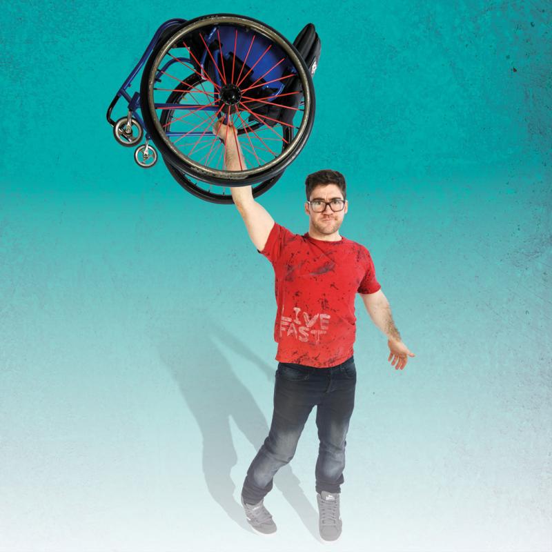 Aaron Simmonds stands against a teal background fading into white. He holds aloft a wheelchair above his head.