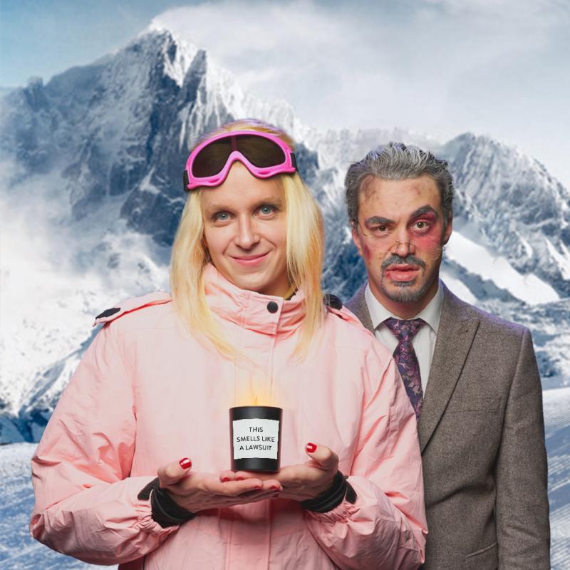 Linus as Gwyneth wears a pink ski suit with long blonde hair and pink ski googles. She holds a GOOP branded candle with the label 'smells like a lawsuit' in front. Directly behind her stands Joseph as Terry who appears bruised, dishevelled and wearing a grey suit and tie for the courtroom. The back drop is a snowy mountain scene.