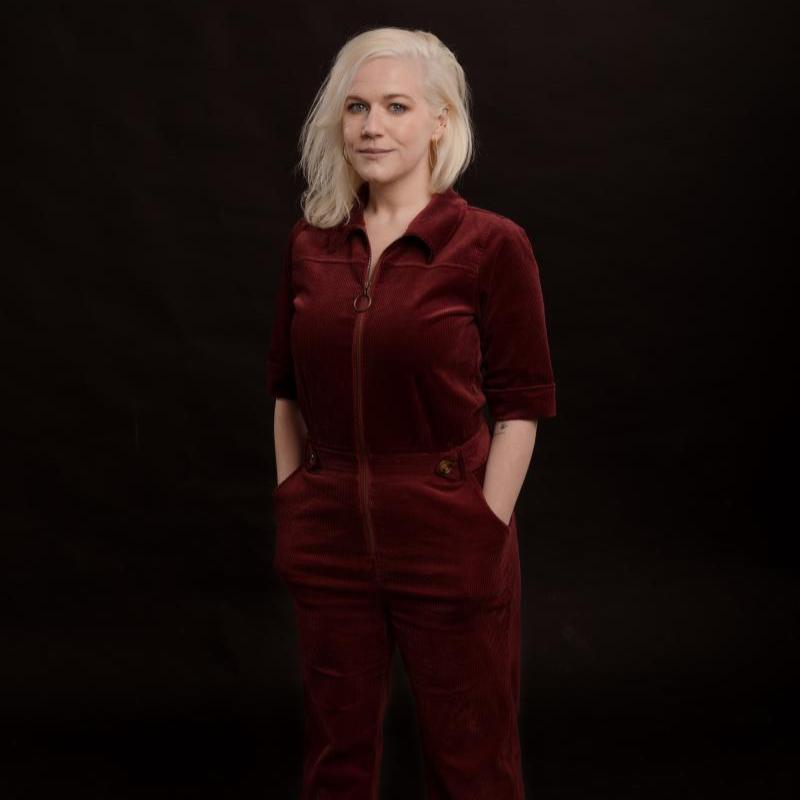 Lulu stands with her hands in her pockets wearing a red velvet jumpsuit against a black background