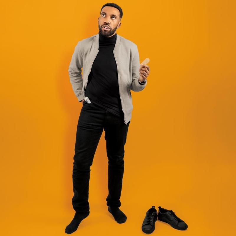 Darran stands against an orange background, one hand in his pocket holding a soggy biscuit. He stands with his shoes off.