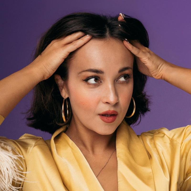 Against a purple background, Sarah Roberts pushes their hair back with their hands. They are wearing a yellow silk shirt with feathered cuffs half out of shop and hoop earrings.