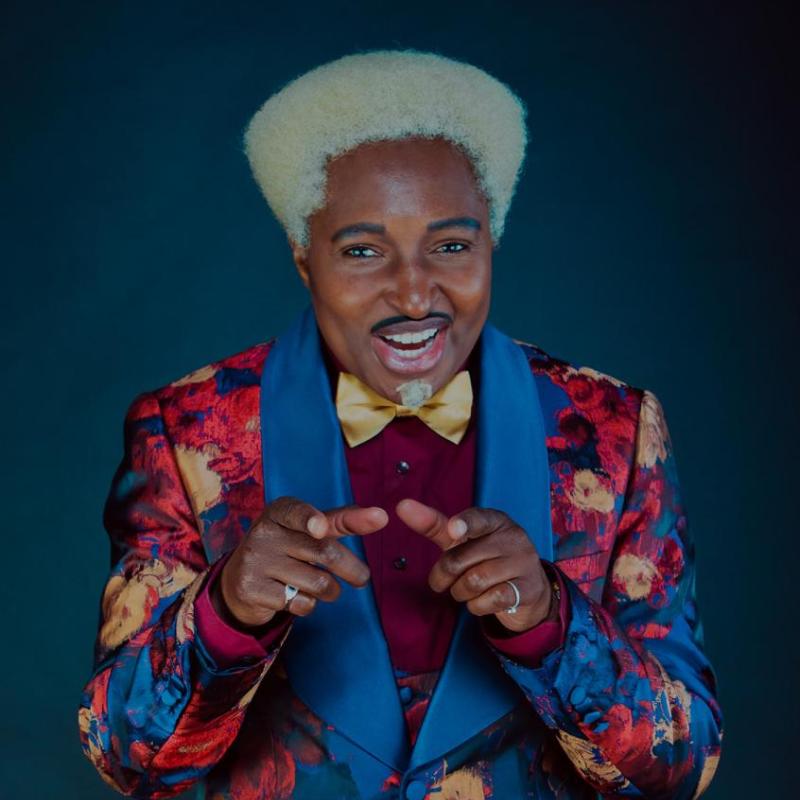 Drag King Don One wears a brightly coloured yellow, red and blue floral suit. They stand in a typical primetime TV host pose with hands cocked, ready to welcome audience members.