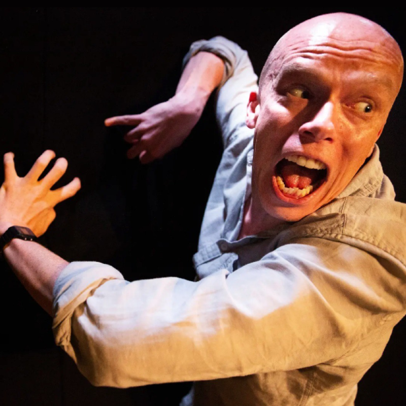 Jordan Brookes' face is close the camera with his eyes wide and mouth open. He wears a beige shirt. His arms are behind him, with his hands tensed.