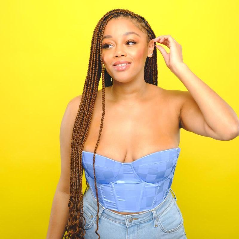 Kyrah wears jeans and a blue strapless corset style top against a bright yellow background. They hold their left hand up to their ear in a relaxed gesture.