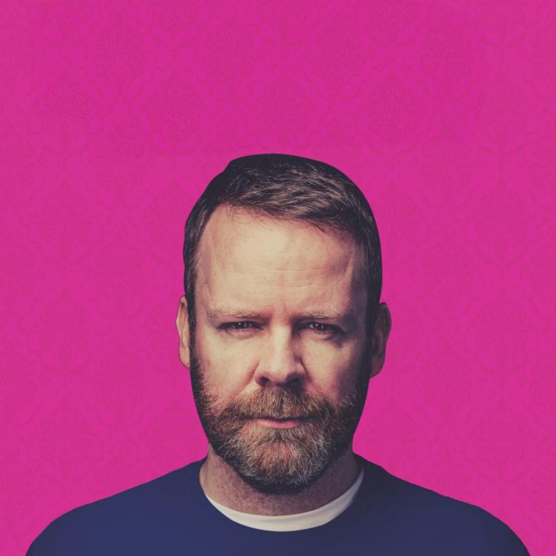 Neil looking at the camera stern-faced against a pink backdrop