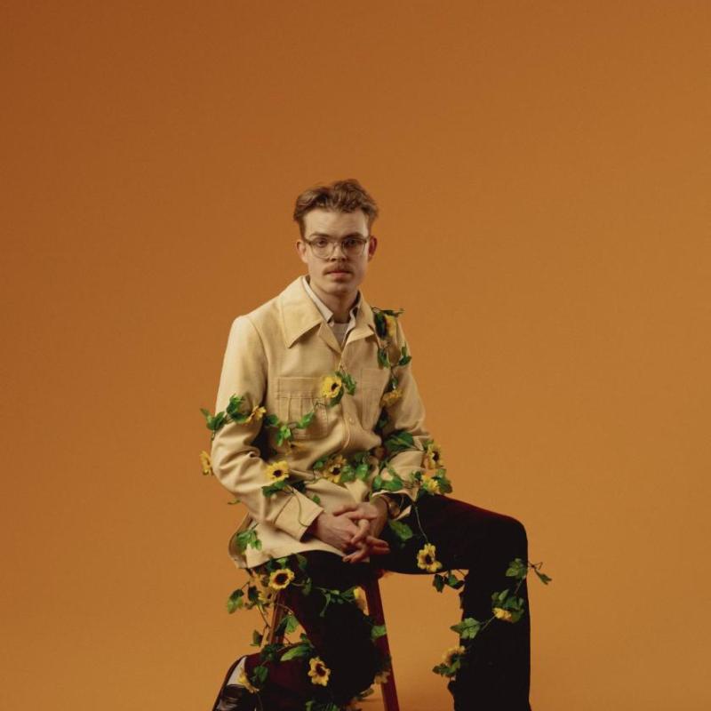 Horatio is sat on a chair in front of an orange background. He is wearing a camel coloured jacket, black tailored trousers and black loafers. There is a vine of sunflowers wrapped around his body.