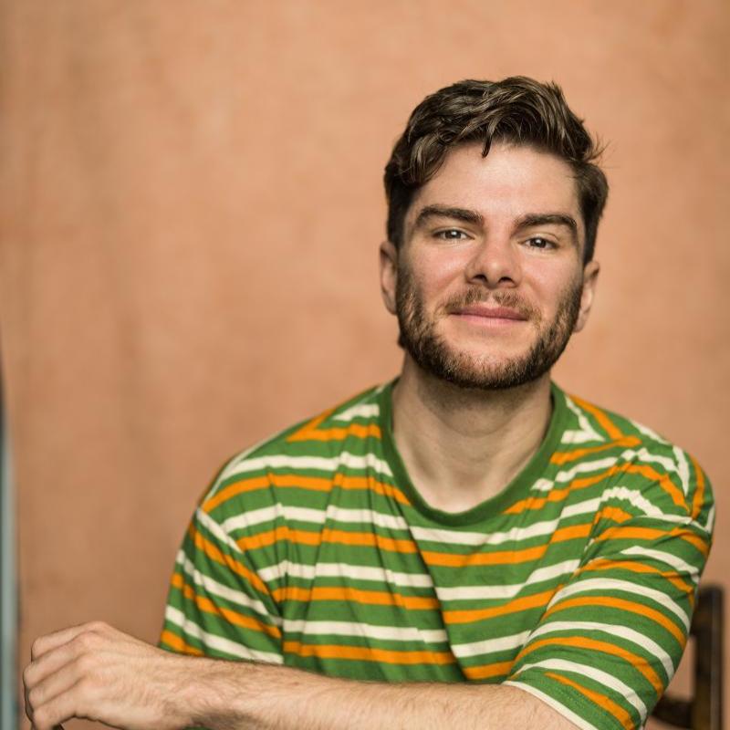 Photo of Christopher wearing a horizontally striped orange, cream and green t-shirt. They look relaxed and are sat on a chair with their arm loosely crossed over their knee.