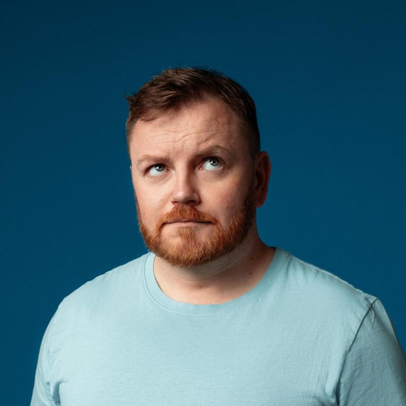 Photo of John looking uncertain with eyes towards the sky. They are wearing a light blue t-shirt against a dark blue teal background.