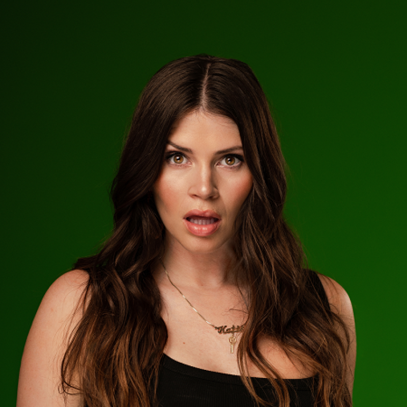 Woman stands in front of green backdrop with her mouth agape in a shocked expression