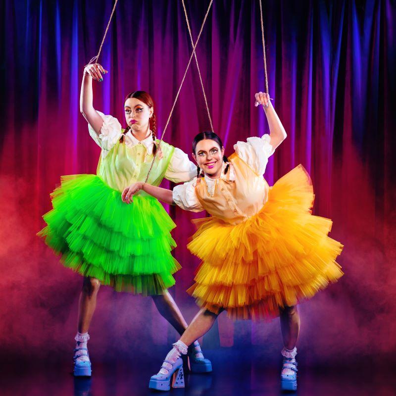 Maddy and Marina are dressed in brightly coloured tutu skirts with platform heels. Their hands are suspended by puppet strings.