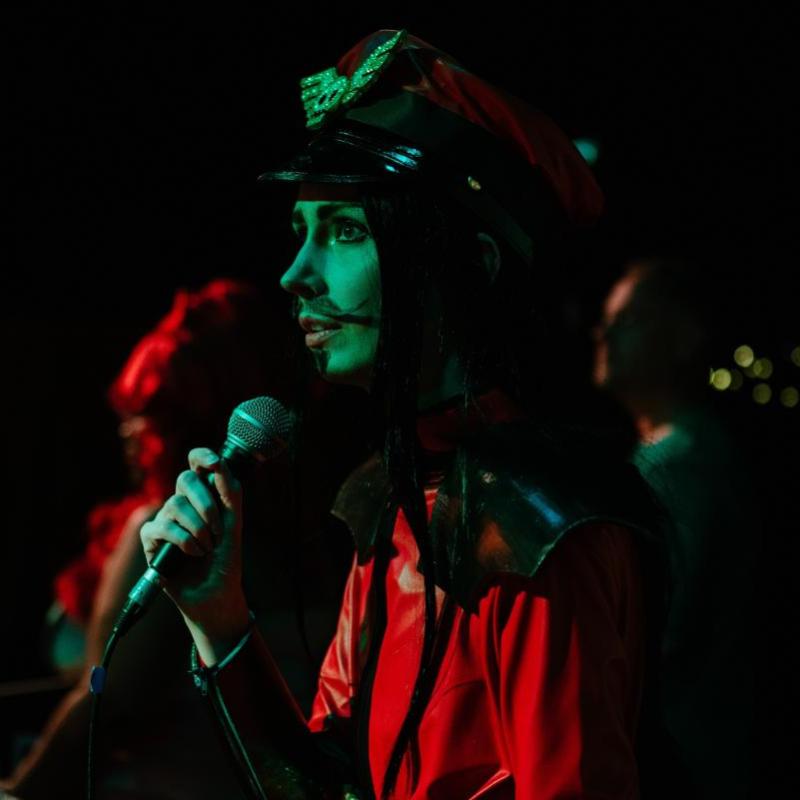 A performer wearing a red ensemble speaks into a microphone around moody, dark, atmospheric lighting