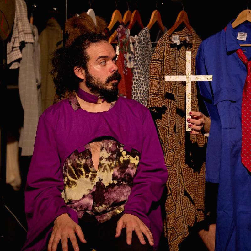 Production shot from Jeezus F*cking Christ. Sergio wears a purple robe sitting down in front of a wardrobe rail. A floating hand appears to their right holding a wooden crucifix.