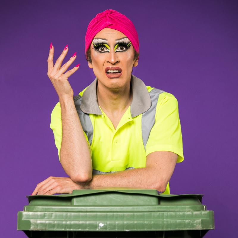 Drag artist wearing a bin collector's clothes and a pink turban. They are resting on top of a green bin with their right hand up and a facial expression that emanates shock and confusion
