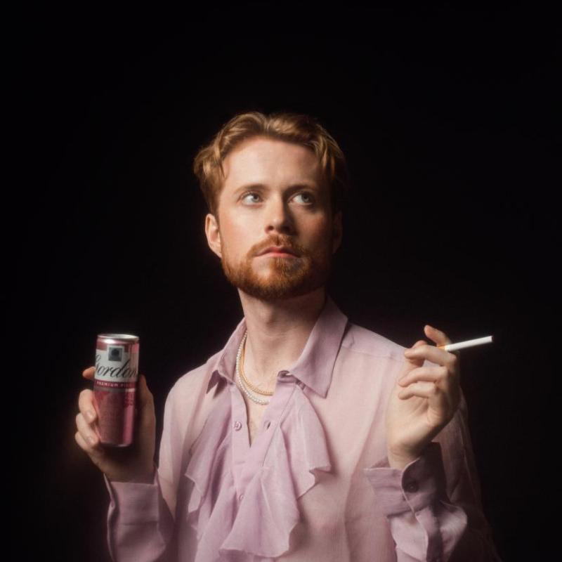 John Tothill wears a flowing, ruffled, pink translucent shirt holding a cigarette and Gordon's Pink Gin & Tonic can in the other.