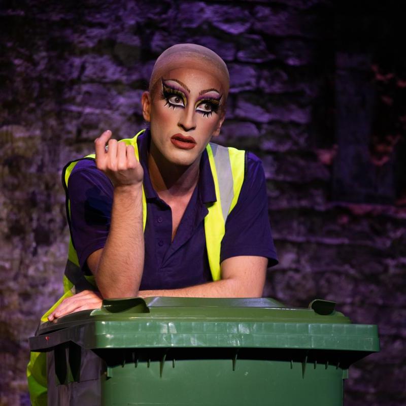 A bin-man leaning on a bin with a full face of drag makeup and a confused expression.