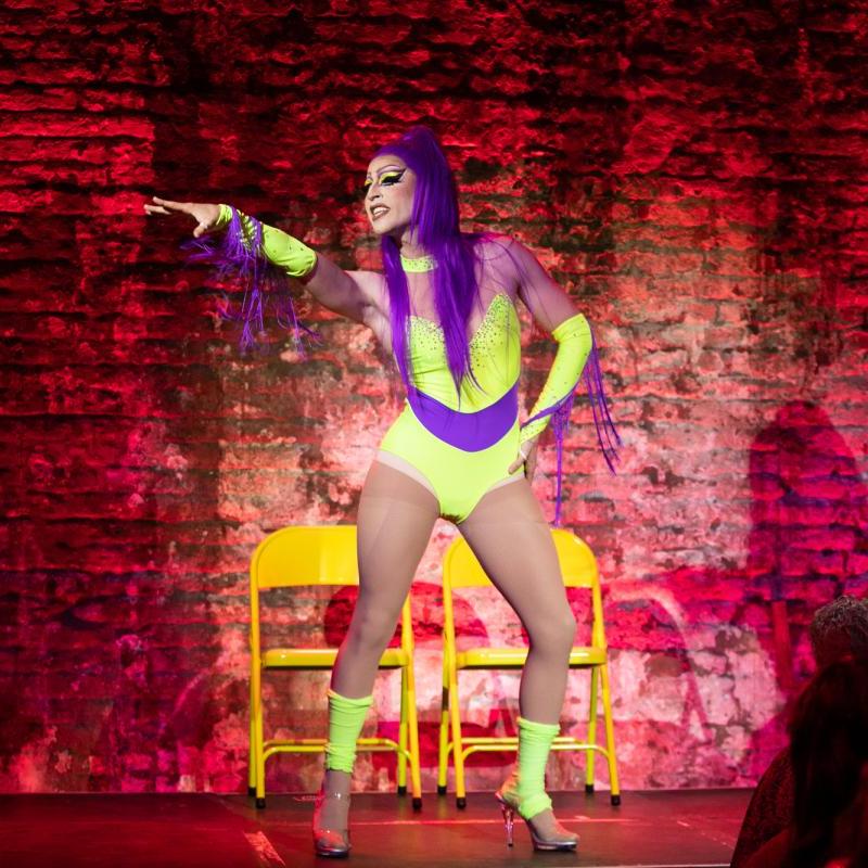 A drag queen in a neon yellow and purple leotard with a purple high ponytail dances against a red backdrop, reaching her right arm out to the audience.