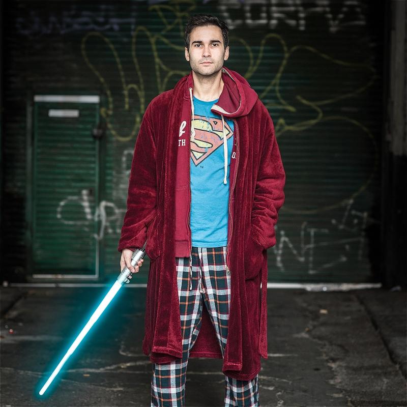 A performer stands facing the camera holding a lightsabre. They wear pyjamas and blue superman t-shirt. 