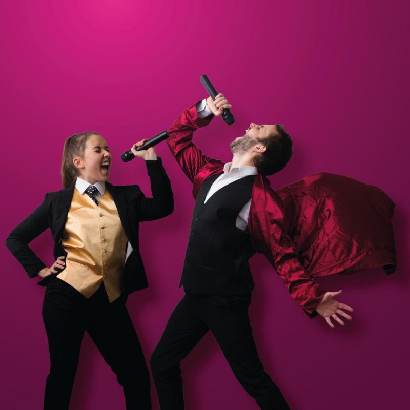 Two performers in waistcoats against a burgundy background singing into microphones