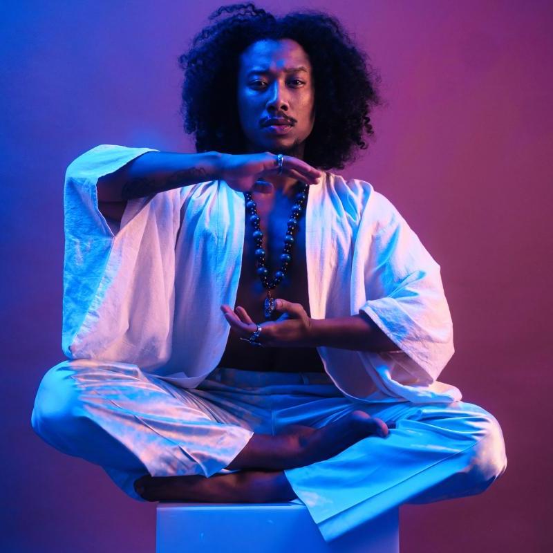 A man sits cross-legged on a white stool in loose white clothing holding his arms to form an energy circle around his chest.