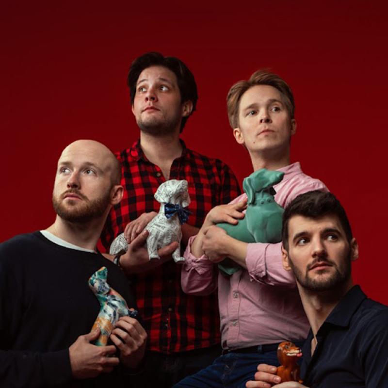 Four performers stand in front of a red backdrop looking in different directions holding toy dogs of varying sizes and colours in a quite gentle way.