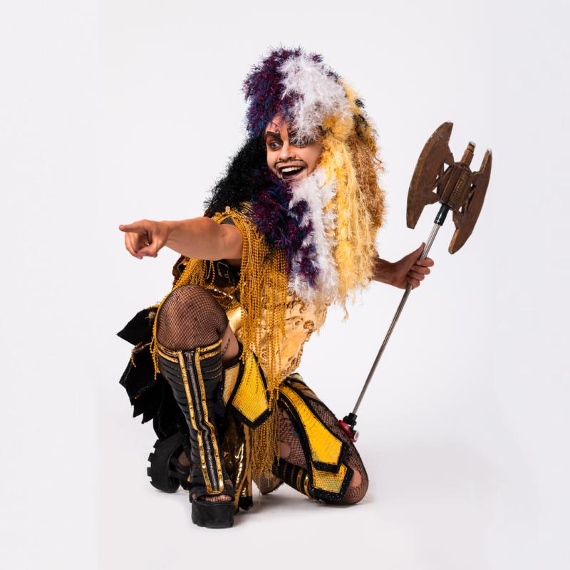 An image of Oedipussi Rex, a white Drag Barbarian with a long beard and wig made out of tinsel in the colours of the Non-Binary Flag. He is Kneeling, his body facing right, while looking and pointing to something out of shot to the left. His expression is gleeful, and he is holding a large double edged axe in his right hand. He is also wearing golden ancient Greek style armour, complete with platform gladiator sandals.