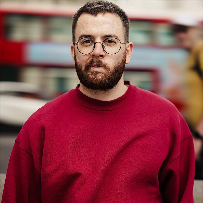 Rich wears a red jumper and looks cheekily at the camera. There is a bus driving by in the background.