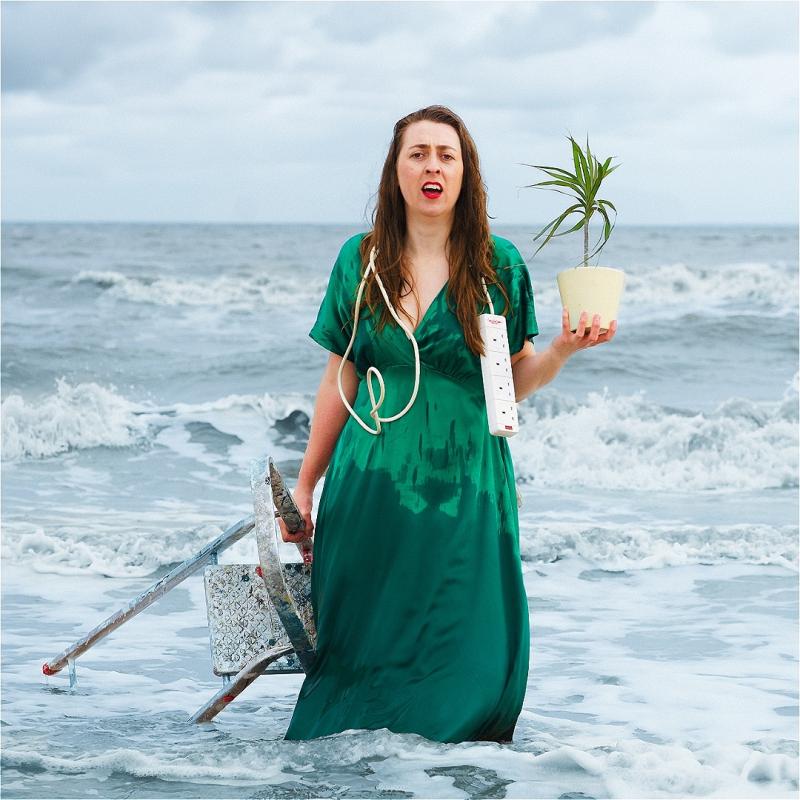 Performer stands in the waves holding a pot plant and ladder with an extension cable draped around their shoulders. The sea spray has left damp splashes all over their green dress.