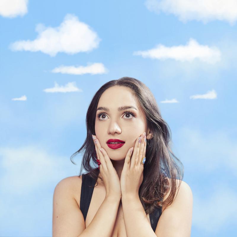 In front of a hazy blue sky backdrop, Bella stands with hands gently cupping their chin. Performer wears bright red lipstick.