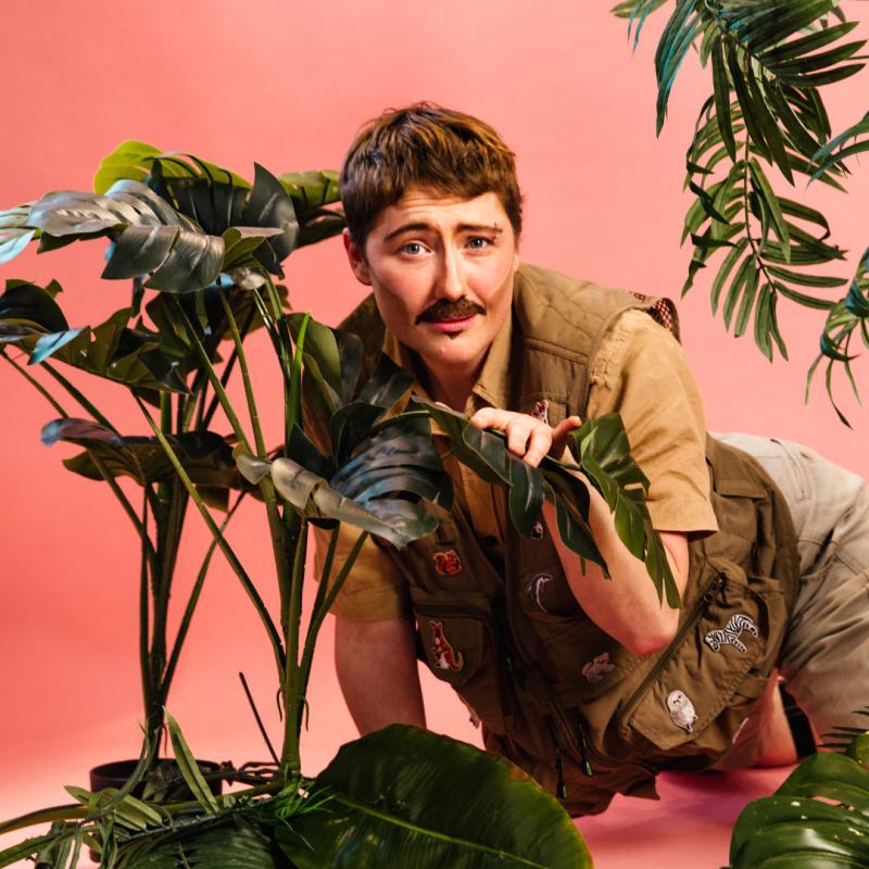 Performer is dressed as explorer in beige crawling through a jungle backdrop of house plants against a pink background.