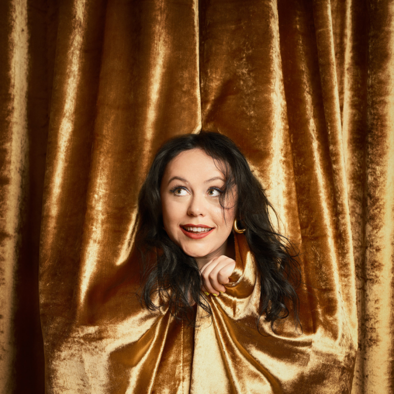 Amy stands amongst a gold velvet curtain looking cheekily out towards the viewer.