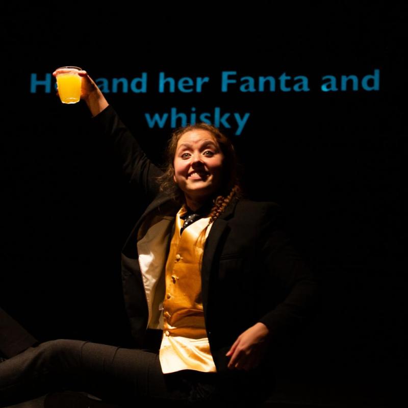 Hannah holds up a glass with an orange liquid with a huge smile on her face. The projection behind says "her and her Fanta and whisky"