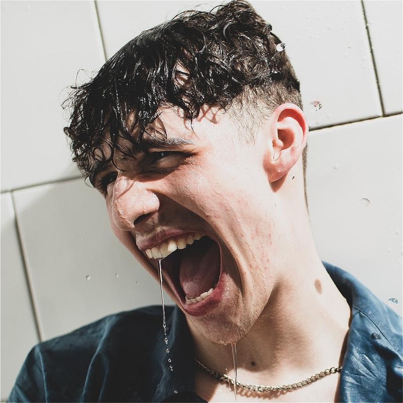 Young man with wet curly hair, wearing a dark shirt, laughing or shouting with joy as water drips from his mouth, against a sunlit white tiled wall.