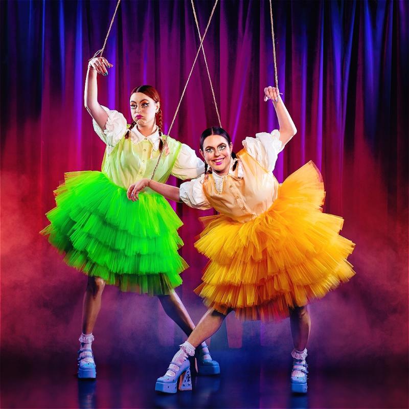 Two performers suspended by strings, resembling marionette puppets, on a stage with a purple curtain background. Both are wearing colorful tutus, the left in green and the right in yellow, with matching suspenders and white socks and high heels. The right performer is smiling at the camera while the left maintains a serious expression.