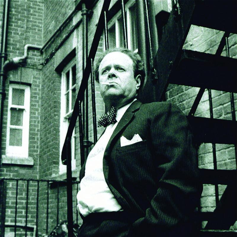Black and white photo of an older man with receding hair, wearing a suit and polka dot bow tie, standing on a fire escape and looking pensively to the side with a cigarette in his mouth, urban building in the background.