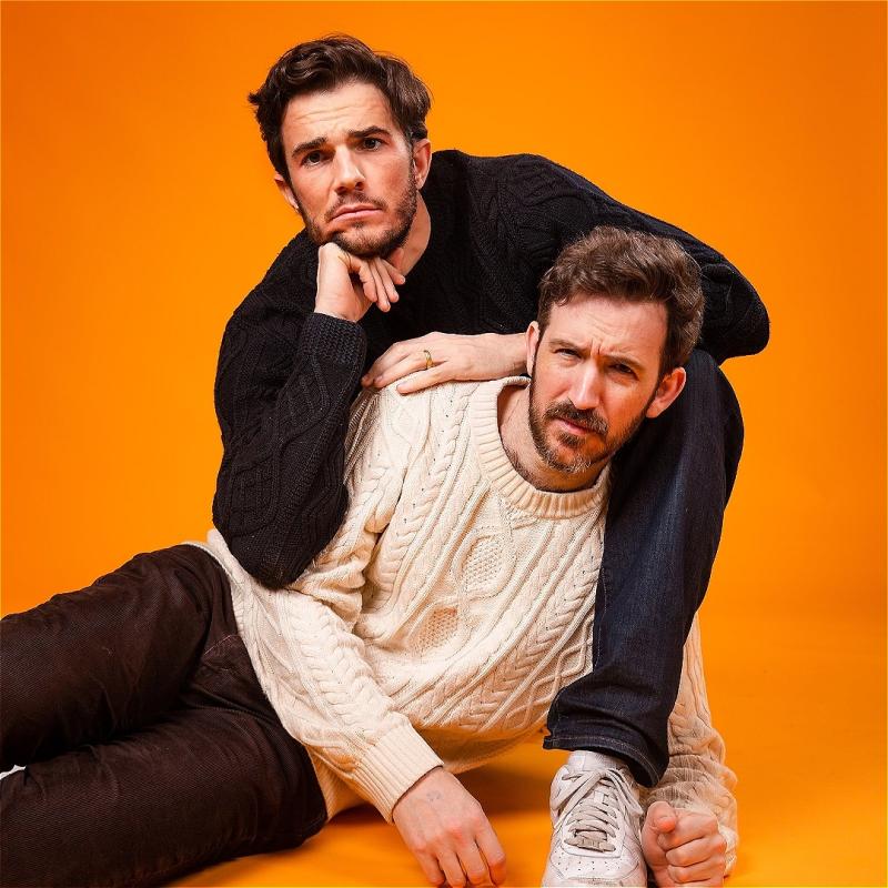 The two performers pose in front of an orange background, with one resting on the other. 