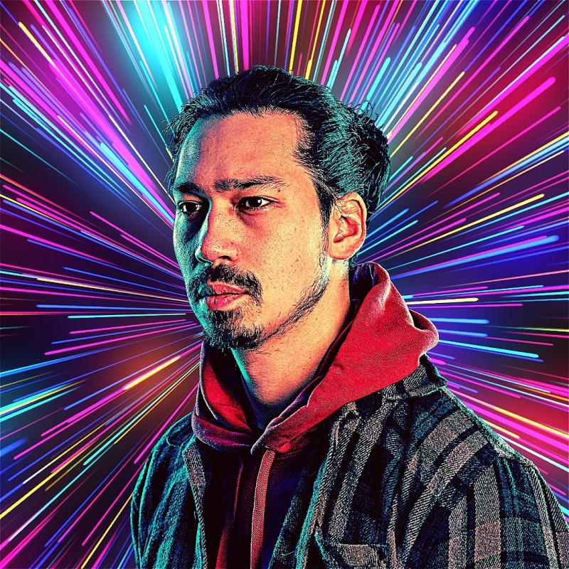 Man with light stubble in a hoodie and jacket, facing slightly left with a serious expression, against a background of vibrant, multicolored light rays emanating from behind him creating a dynamic and futuristic effect.