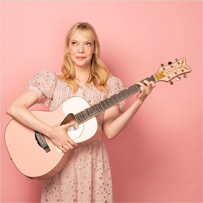 Woman with blonde hair in a pink lace dress smiling and playing a pink acoustic guitar against a pink background.
